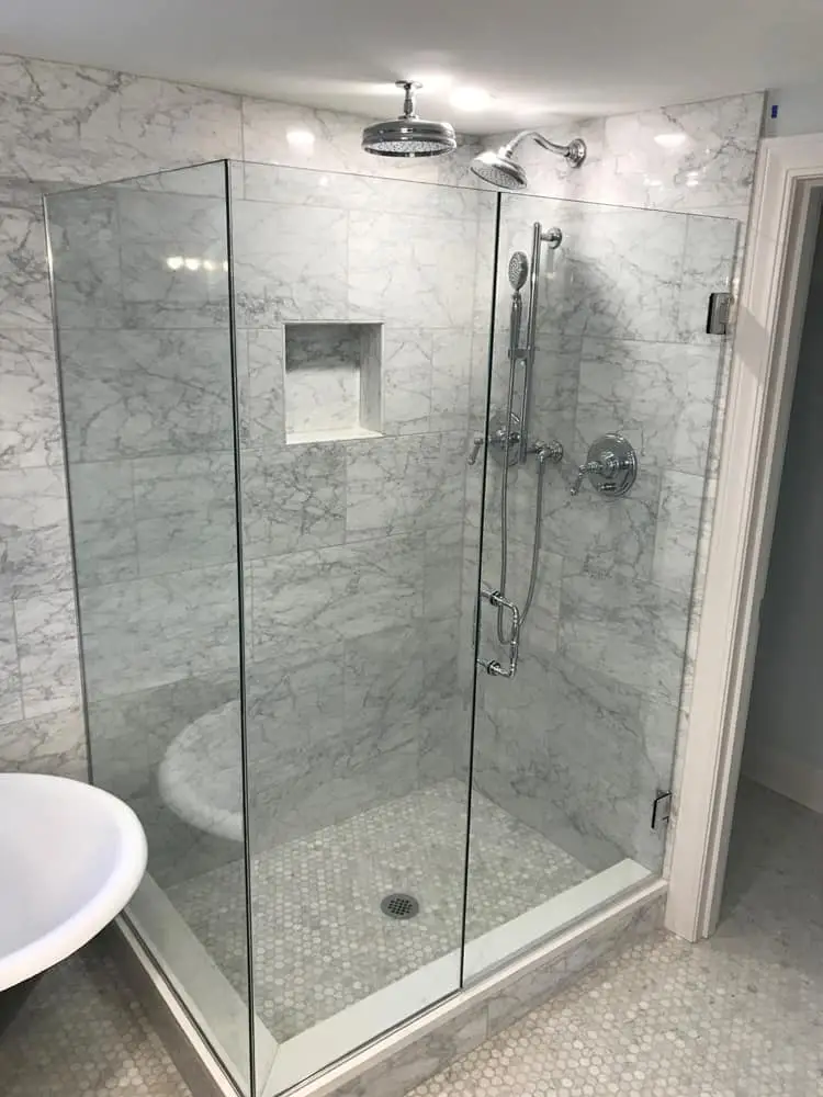 Frameless 90-degree corner shower enclosure with a hinged door and chrome hardware in a Carrara marble bathroom.