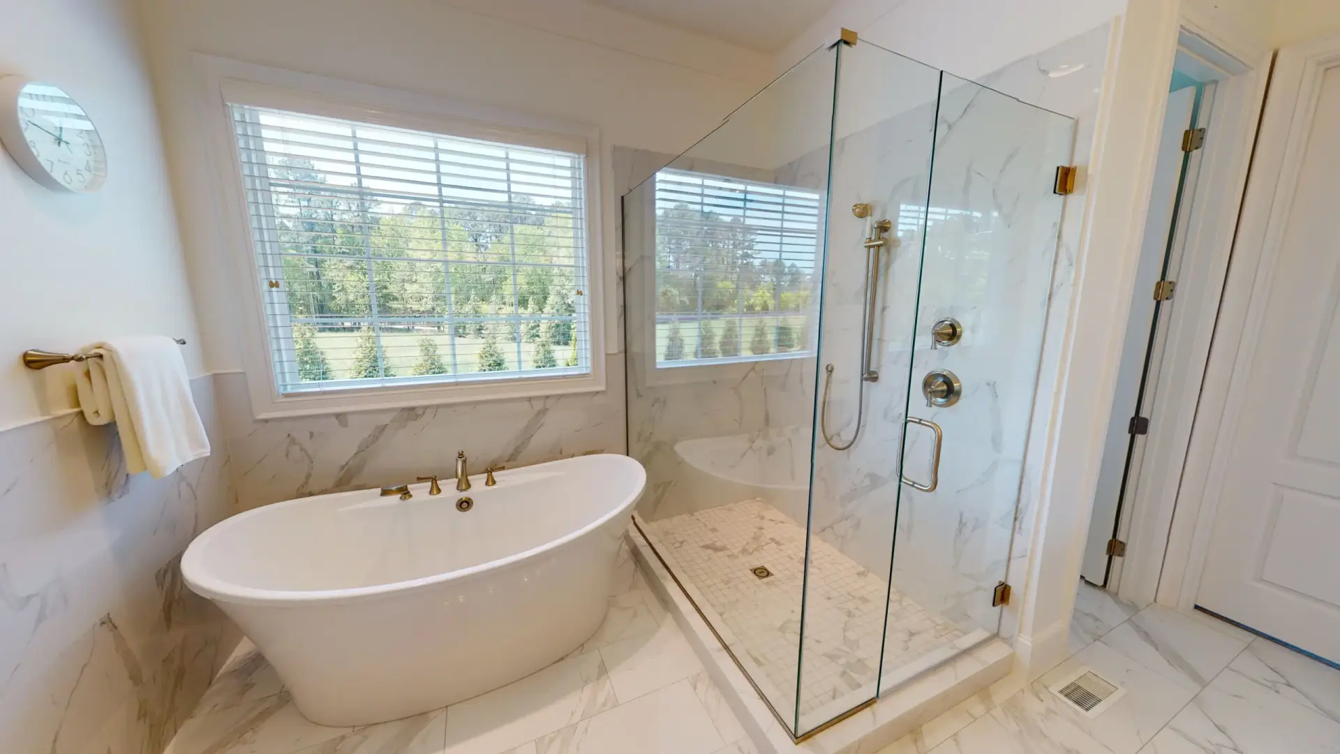Frameless 90-degree shower with hinged door and satin brass hardware in a sunlit bathroom with a freestanding tub.