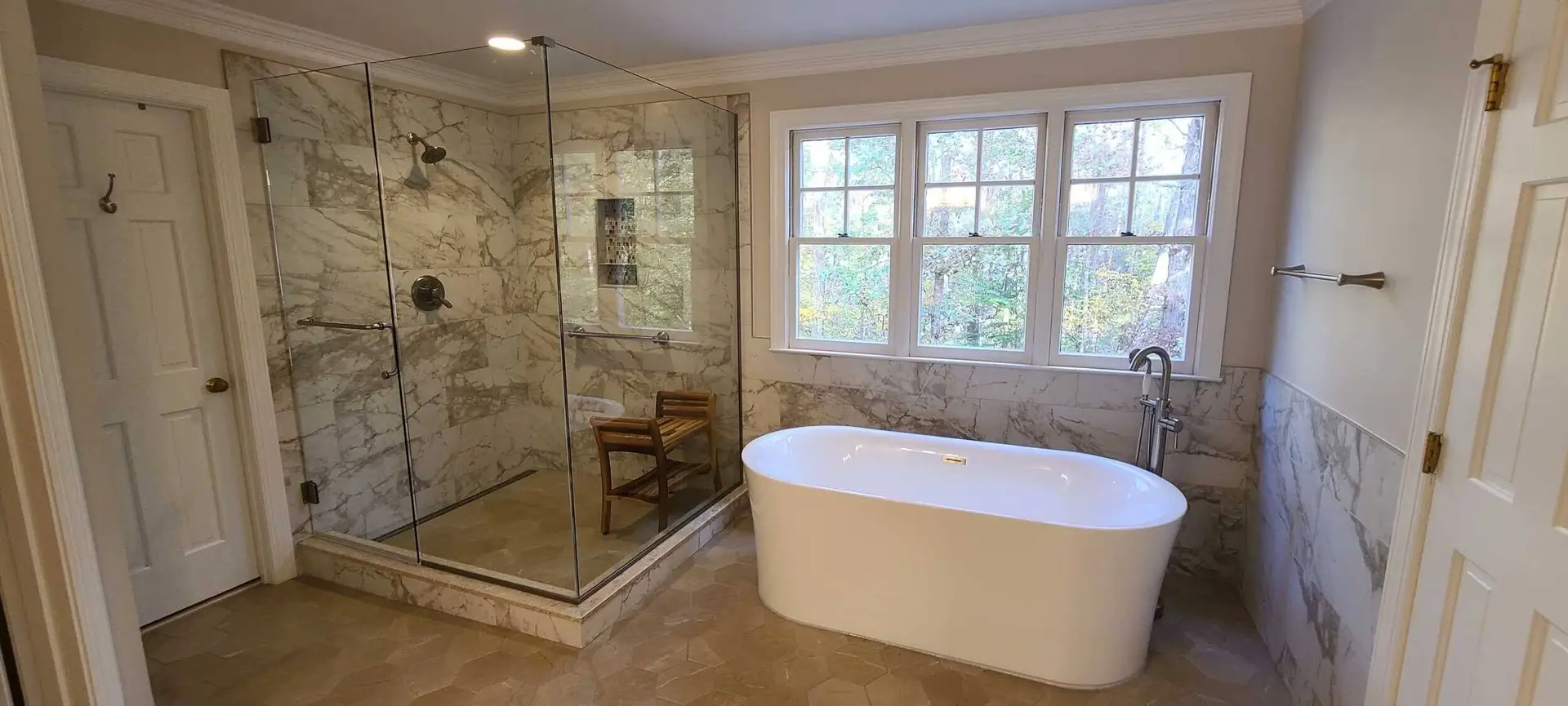 Inline frameless shower with two glass doors and fixed panels, oil-rubbed bronze hardware, and floor-to-ceiling marble surrounding a sunlit window.