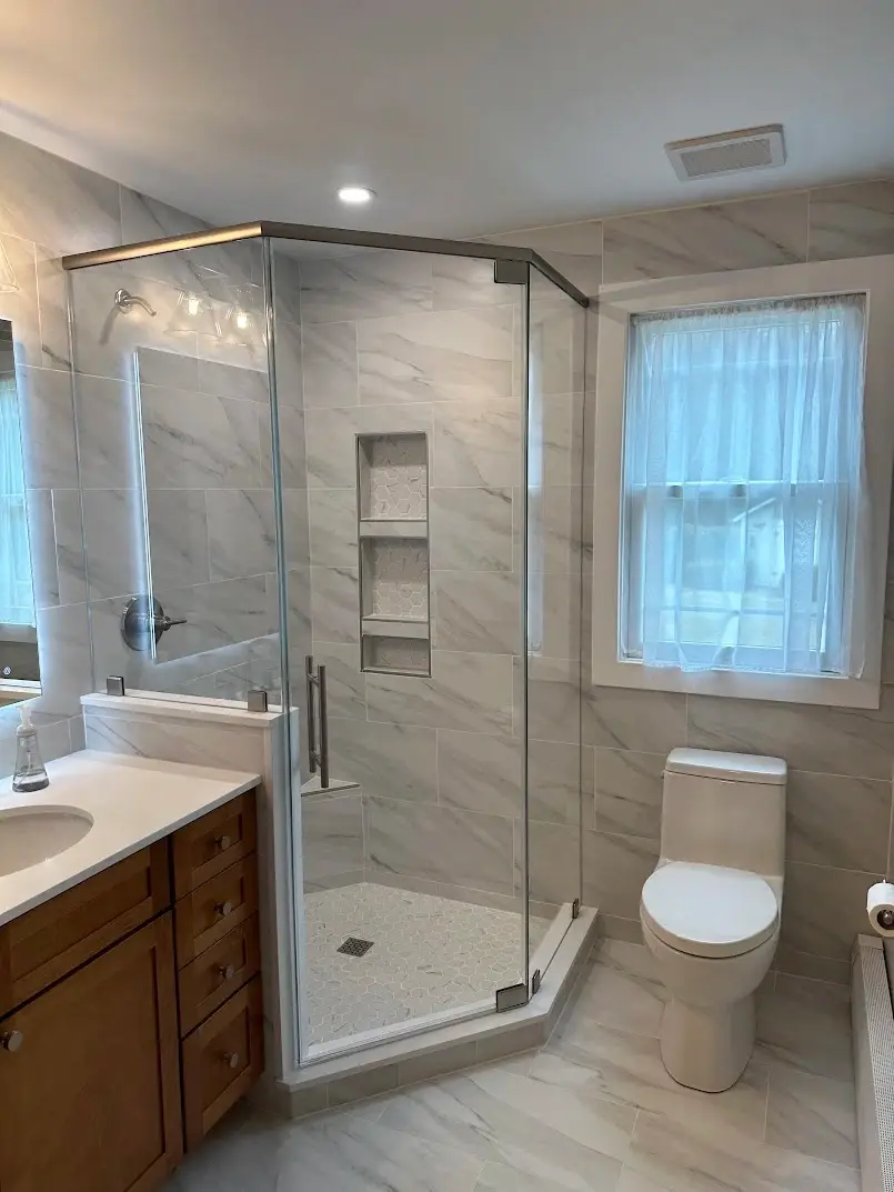 Compact neo-angle shower enclosure with brushed nickel hardware, marble-look porcelain tile, and a honey oak vanity.