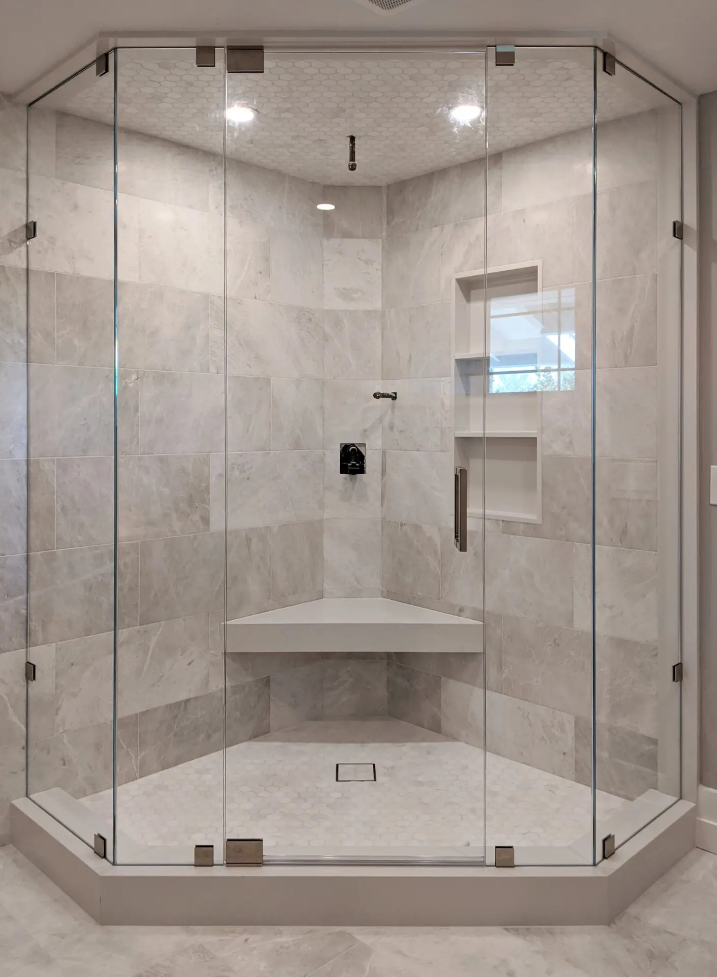 Tall frameless neo-angle shower enclosure with brushed nickel hardware, grey marble walls, hex mosaic ceiling detail, and a floating corner bench.