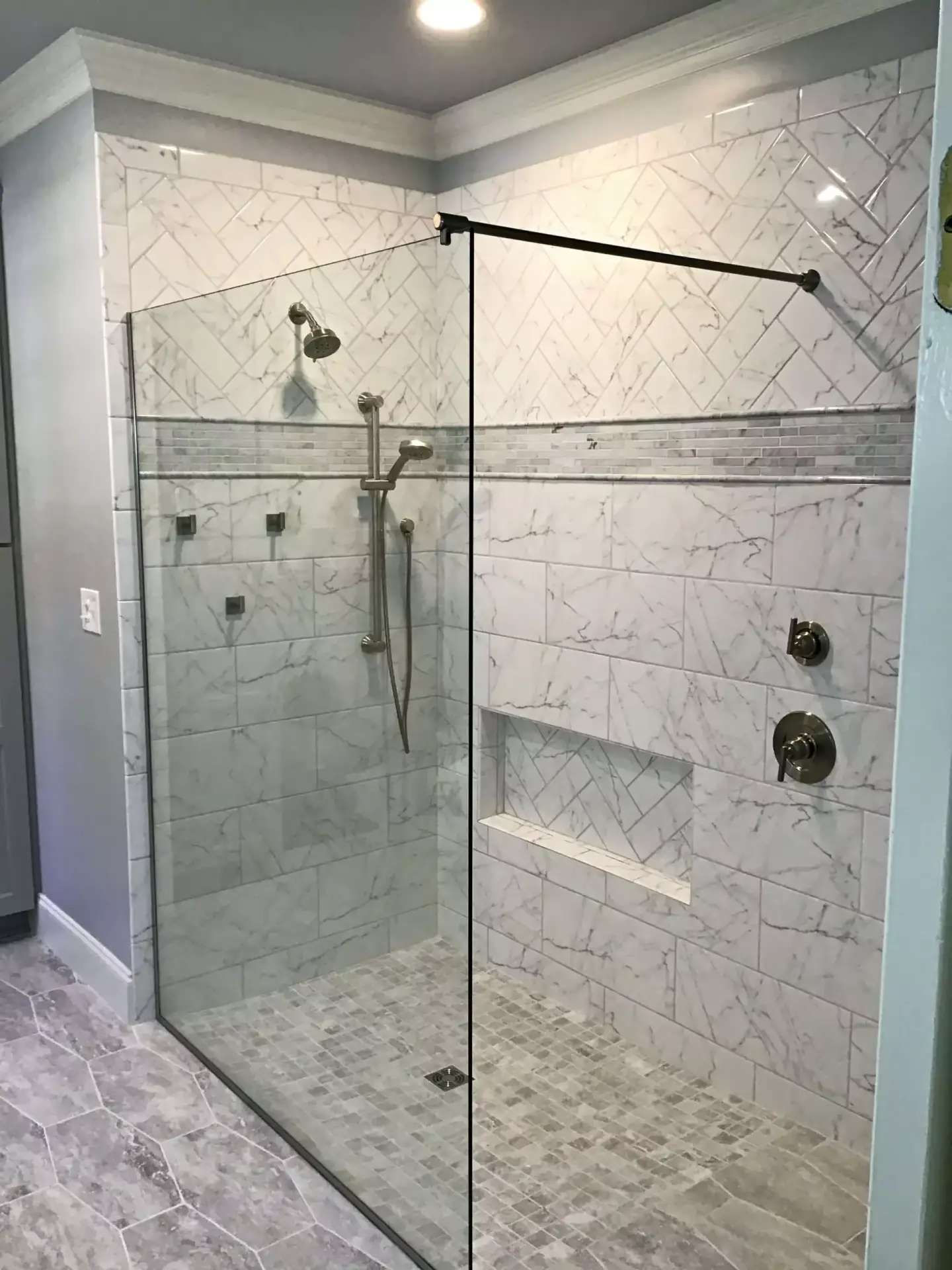 Open walk-in shower with two fixed glass panels meeting at a 90-degree corner and herringbone marble walls.